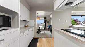 A modern kitchen with white cabinetry, a black microwave, and a wooden floor leading to a dining area with a view. - West Nine Stunning Views & Hot Tub (Port Angeles)