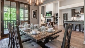 A dining area with a wooden table set for six, a chandelier, and a view of the outdoors through large windows.