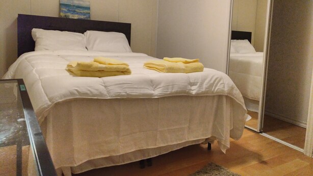 A neatly made bed with white linens and yellow towels, a painting on the wall, and a mirror reflecting the room. - Toronto vacation home in North York with free parking (minimum 28 days stay)  (North York)