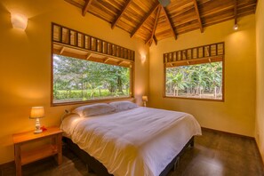 Un dormitorio con techo de madera, dos ventanas amplias que dan a zonas verdes, una cama con sábanas blancas y una mesita de noche con lámpara.