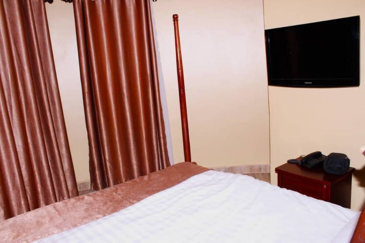 Habitación de hotel con cama, cortinas, una barra de madera, televisor en la pared y mesita de noche con teléfono y bolso.