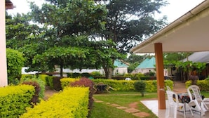 A covered outdoor area with a lawn, yellow flowering bushes, and a table with chairs. - Lyden Cottages (Kampala)