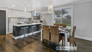 A modern kitchen with white cabinets, a central island with bar stools, and a dining area with wicker chairs. - Settle Inn, Settlement Rd holiday house, close to shops and beach! (Cowes)