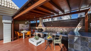 An outdoor patio with a wooden deck, a barbecue grill, a dining table with chairs, and a decorative wall. - Protaras Villa Rocco Near The Beach (Protaras)