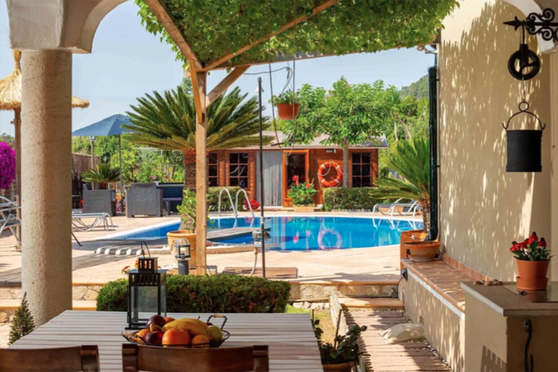 Uma área de piscina com vista para uma casa, uma mesa com frutas e um lança-luz pendurado.
