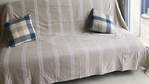 A room with a striped sofa, two pillows, a framed picture of a sailboat, and a window with a view of a blue building. - PORT CROUESTY apartment with pool (Arzon)