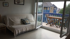 A room with a sofa, a small table, and a balcony with a view of houses. - PORT CROUESTY apartment with pool (Arzon)
