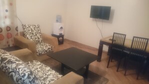 A living room with a sofa, a patterned armchair, a coffee table, a TV mounted on the wall, and a small table with chairs.