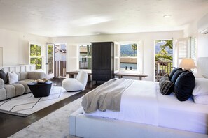 A modern bedroom with a bed, sofa, and desk, large windows with shutters, and a view of greenery.