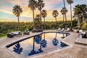A swimming pool with lounge chairs and a patio area, surrounded by palm trees and a sunset view.