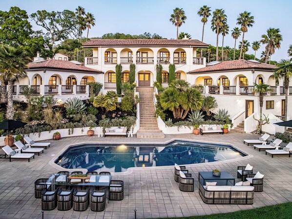 A large building with multiple balconies, a central staircase, a swimming pool, and outdoor seating areas.