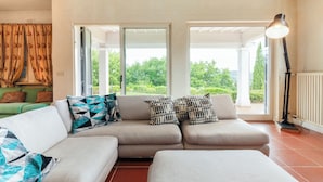 A modern living room with a beige sofa, patterned cushions, and a view of greenery through large sliding doors. - Villa Ninfeo 10+4, Emma Villas (Arezzo)