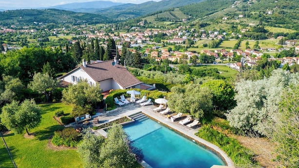 A large, curved swimming pool surrounded by a lawn and trees, with a villa and lounge chairs in the foreground. - Villa Ninfeo 10+4, Emma Villas (Arezzo)