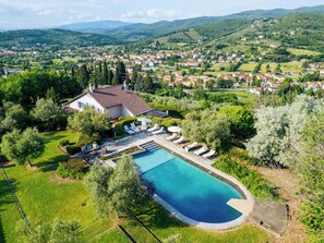 Une grande piscine incurvée, entourée d’une pelouse et d’arbres, avec une villa et des chaises longues au premier plan.