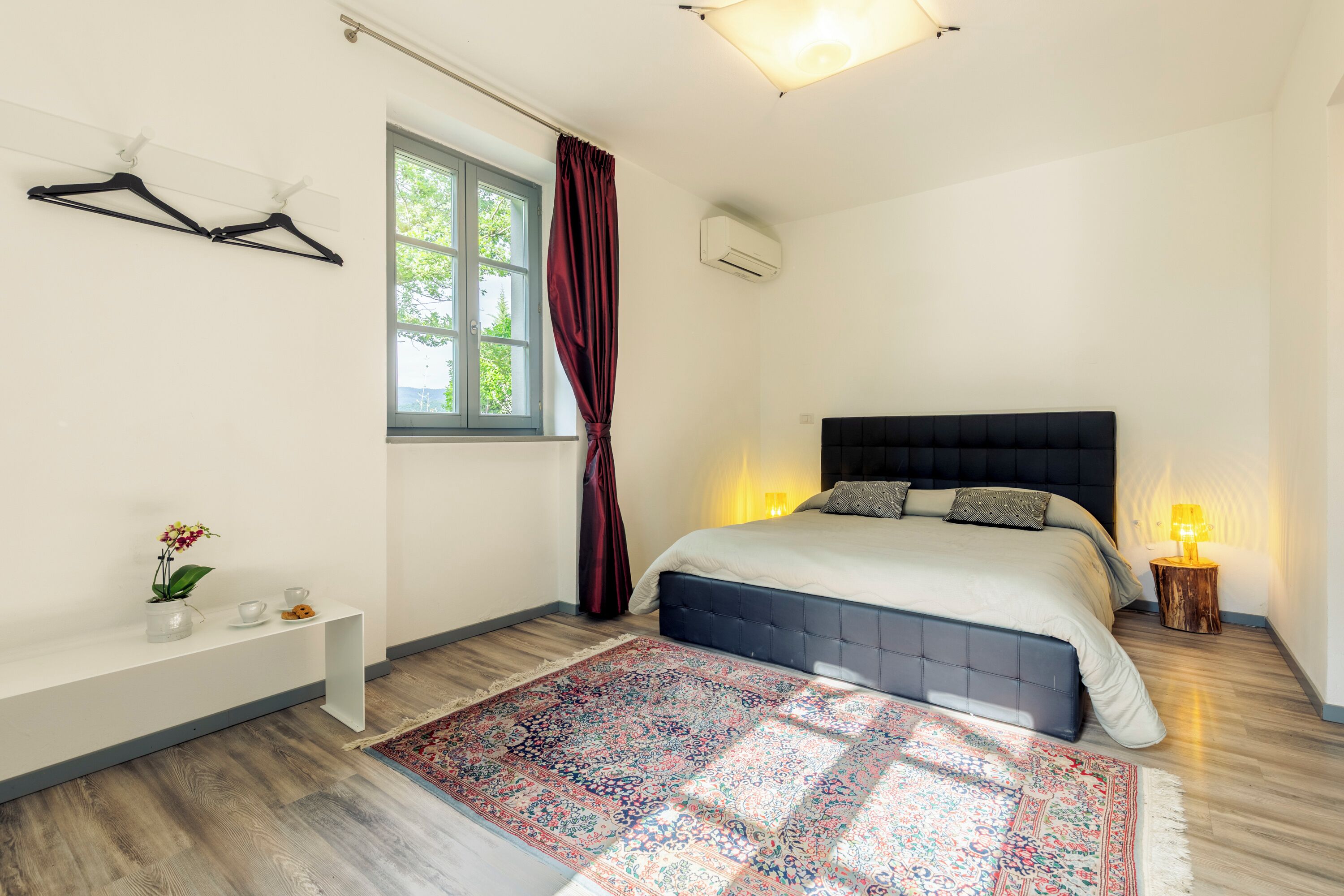 Une chambre à coucher comprenant un lit, une table de chevet, une fenêtre avec des rideaux, un appareil de climatisation et un tapis à motifs.
