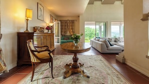 A spacious living area with a round wooden table, a sofa, and a chair. There is a floral rug on the floor, and a cabinet is visible in the background. - Villa Ninfeo 10+4, Emma Villas (Arezzo)