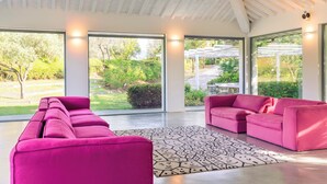 A spacious living area with a modern design, featuring a long pink sofa and a smaller pink armchair, large windows offering a view of the garden, and a patterned rug on the floor. - Villa Ninfeo 10+4, Emma Villas (Arezzo)