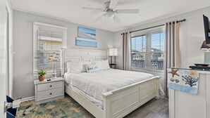 A bedroom with a white bed, a ceiling fan, a nightstand with a lamp, and a view of the city through large windows. - Ce Chell, Spacious 4 BR Channel Home with Beautiful Channel Views and Floating Dock (NORTH MYRTLE BEACH)