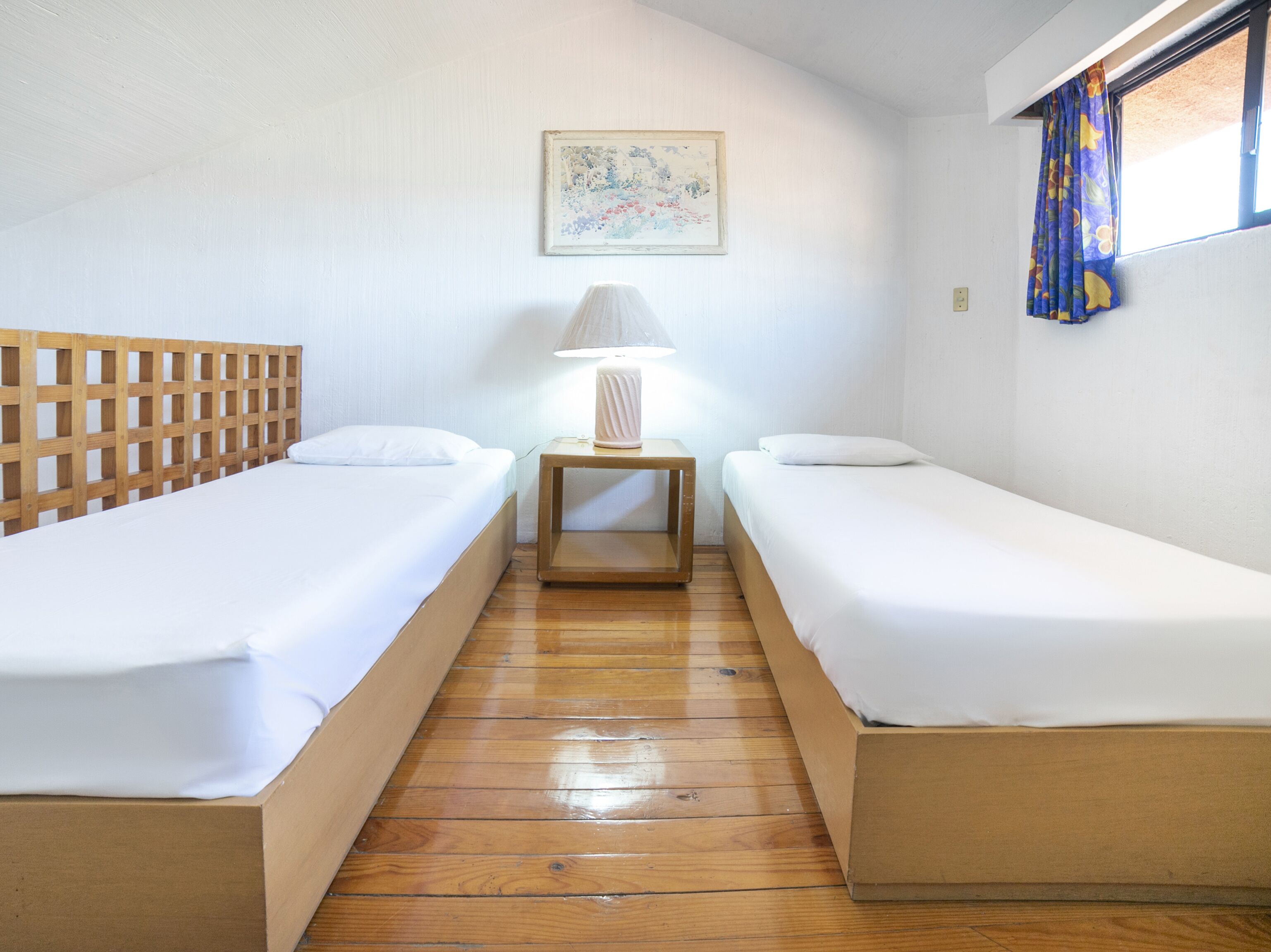 Una habitación pequeña y limpia con dos camas individuales, cabecera de madera, mesita de noche con lámpara y ventana con cortinas.