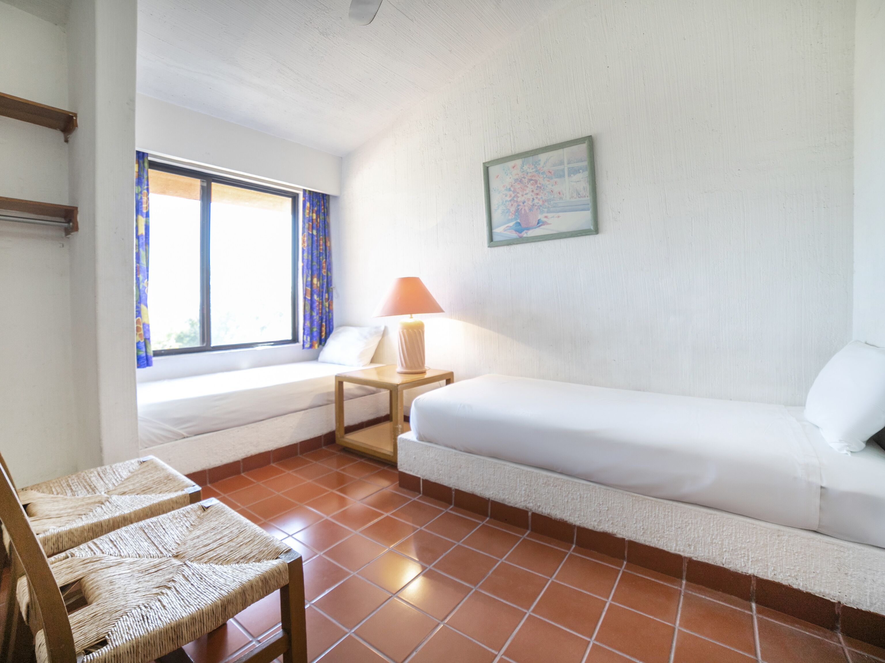 Una habitación con una cama, una silla, una mesa con una lámpara, una ventana con cortinas y un cuadro en la pared.