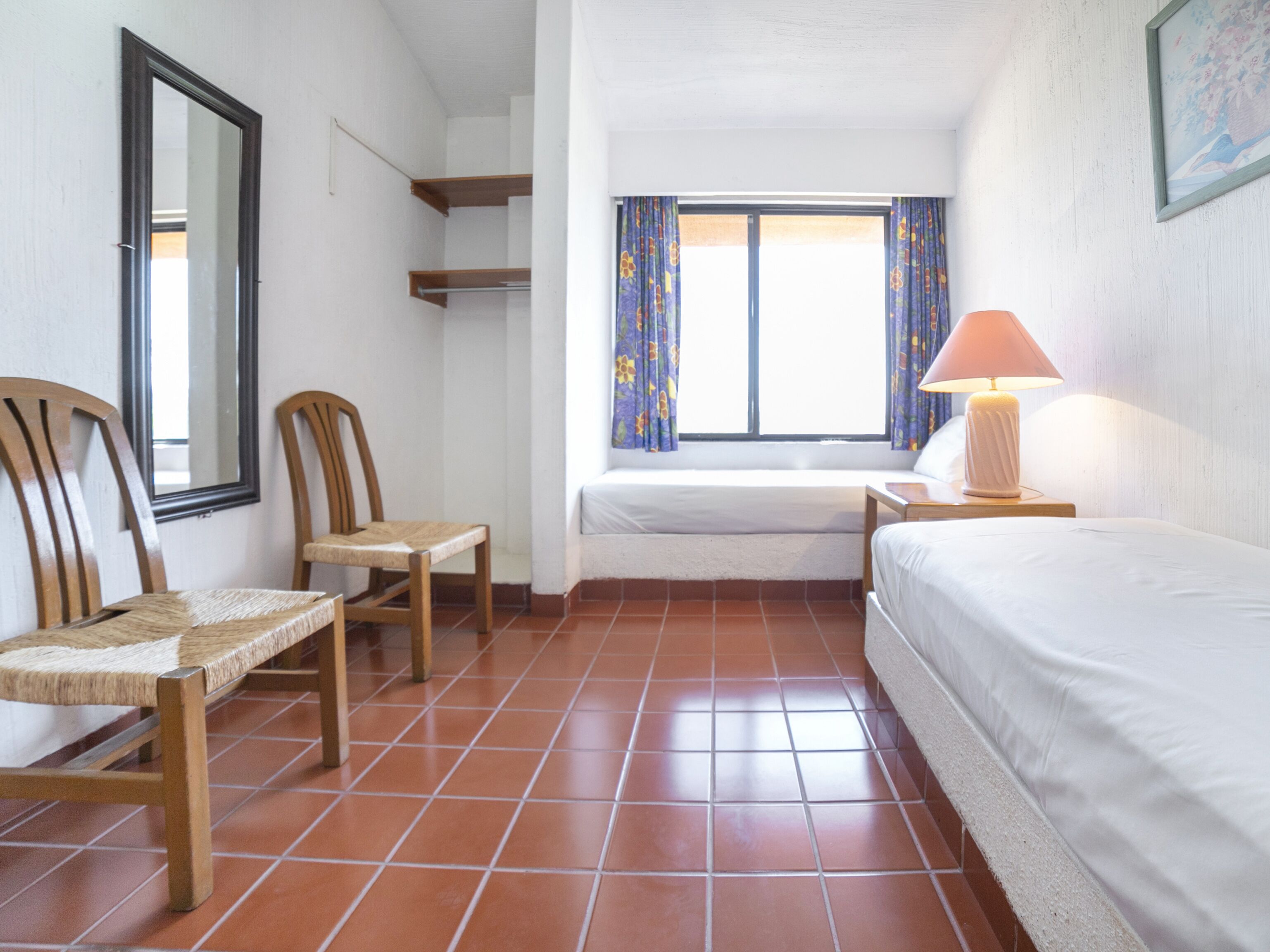 A small, clean room with a bed, two chairs, a nightstand, a lamp, and a window with curtains.