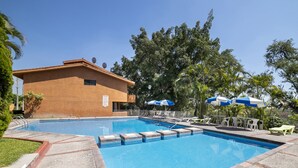 Una piscina con borde de baldosas, rodeada de sillas de descanso y sombrillas, y un edificio con antenas parabólicas al fondo.