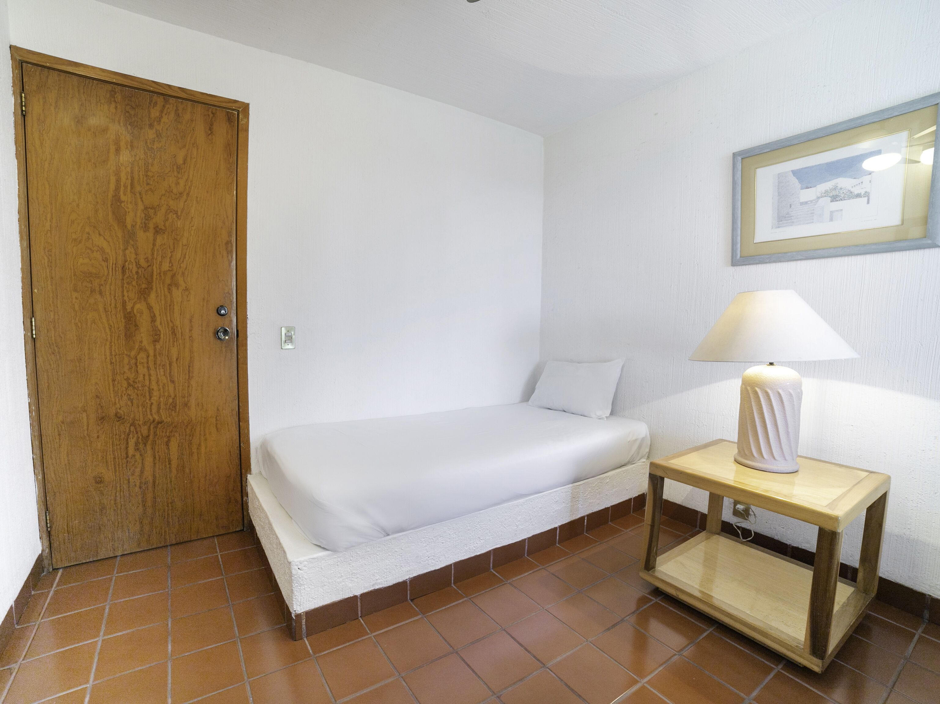 A small, clean room with a single bed, a wooden door, a small table with a lamp, and a framed picture on the wall.