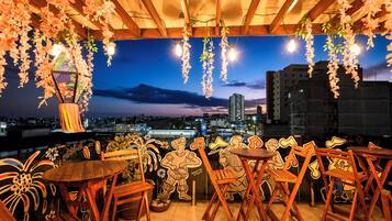 Un área de estar al aire libre con mesas y sillas de madera, adornos florales colgantes y vistas de la ciudad al anochecer.