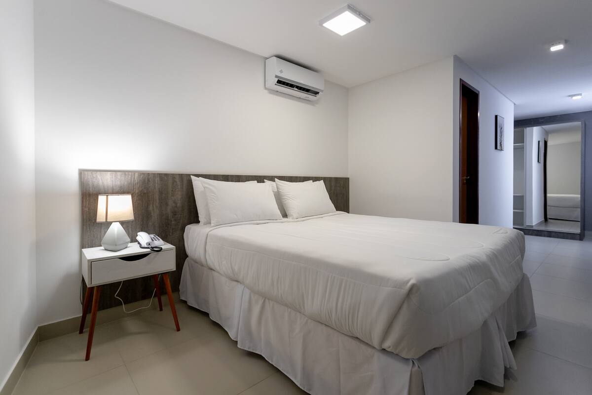 A hotel room with a large bed, a bedside table with a lamp, and a wall-mounted air conditioner.