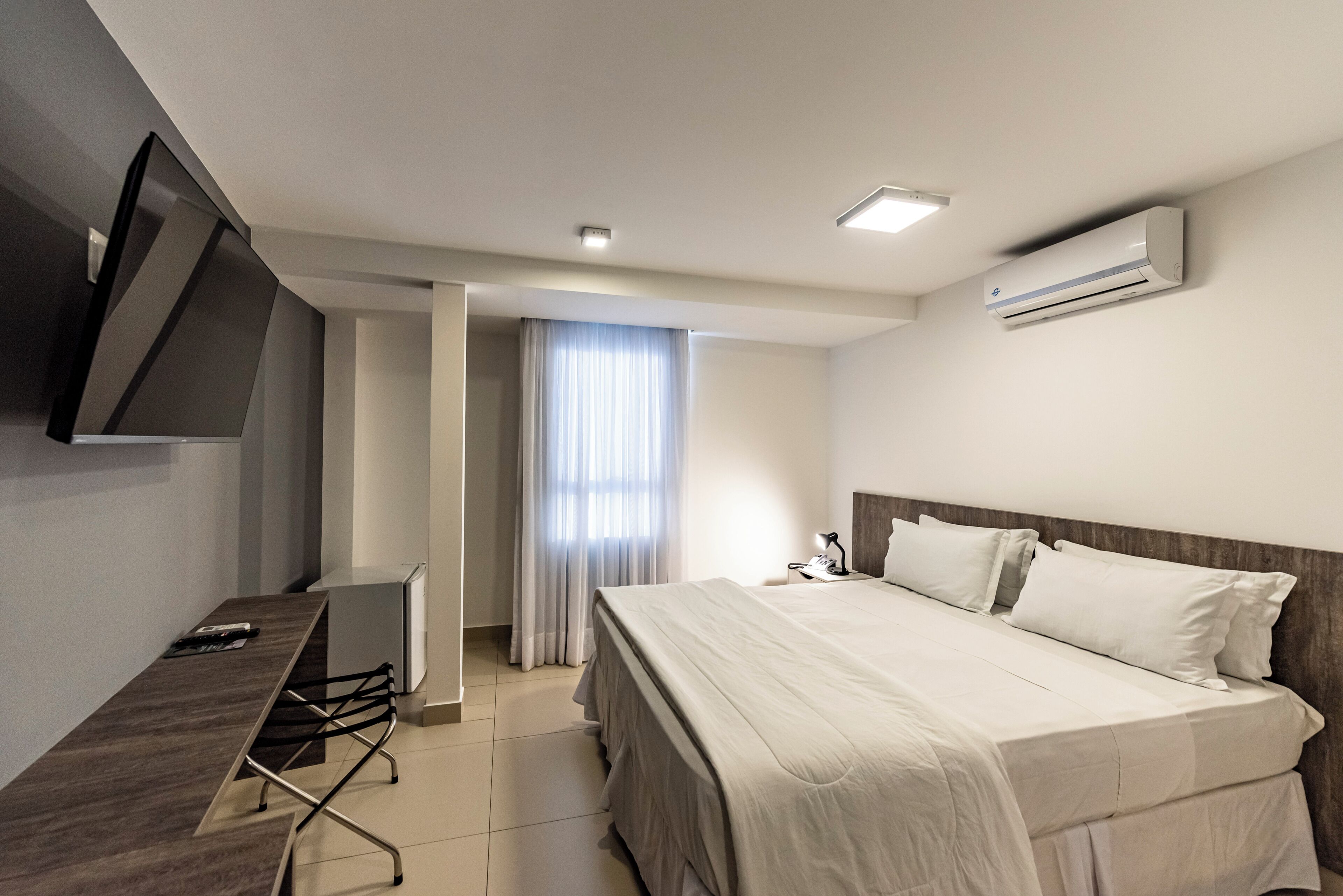 A hotel room with a bed, a desk, a chair, a TV, and an air conditioning unit.