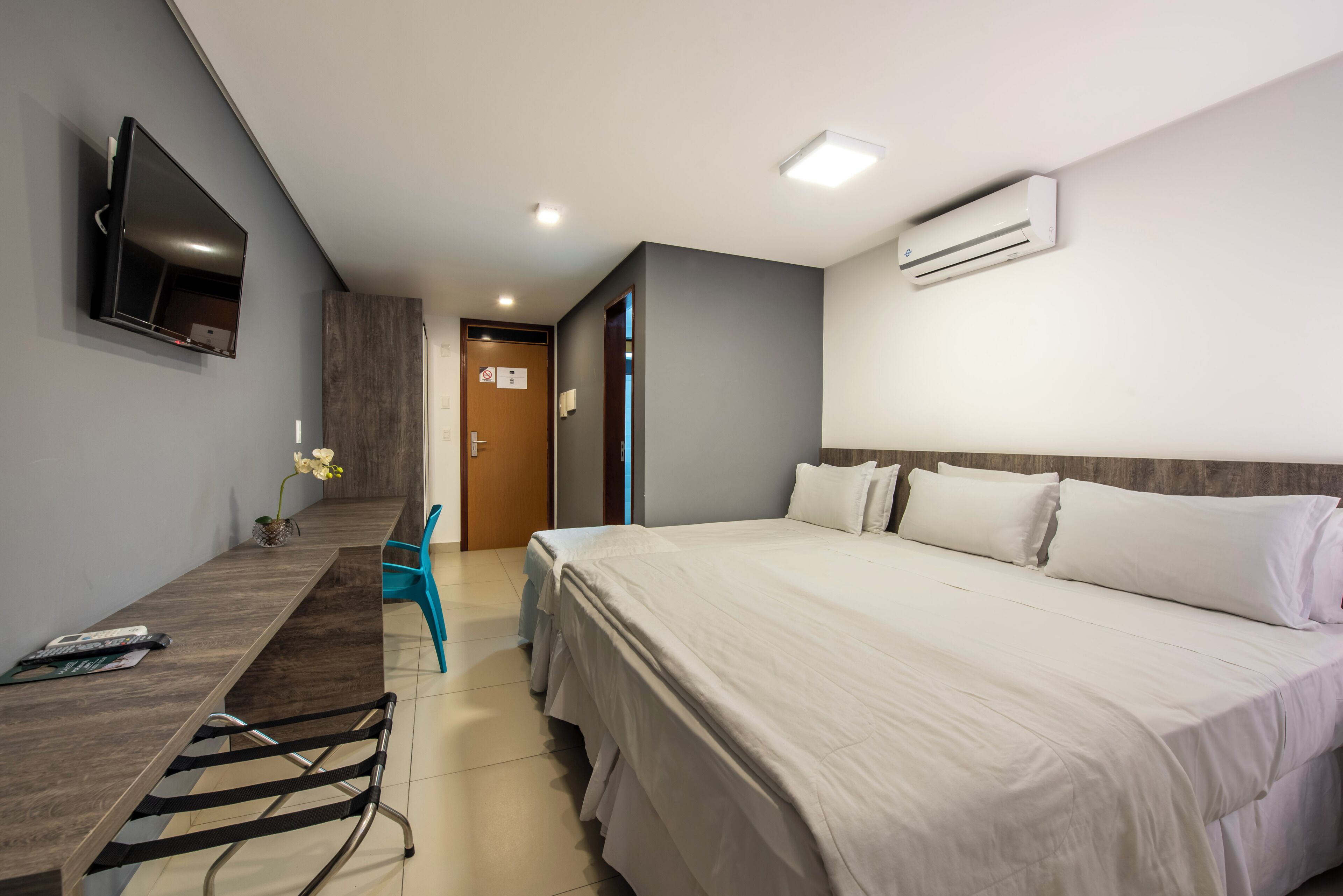 A hotel room with a bed, a desk, a chair, a TV, and an air conditioning unit.