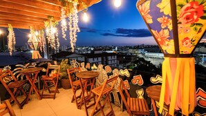 Un espacio al aire libre con sillas y mesas de madera, decoración floral colgante y vistas a un paisaje urbano al atardecer.