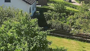 Un quartier résidentiel avec une maison blanche, un petit abri de jardin et un jardin bien entretenu, agrémenté de haies et d’un sentier.