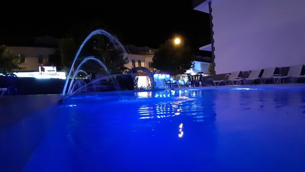 A nighttime swimming pool with water fountains, lounge chairs, and a building in the background. - Moonlight Apart Hotel (Ortaca)
