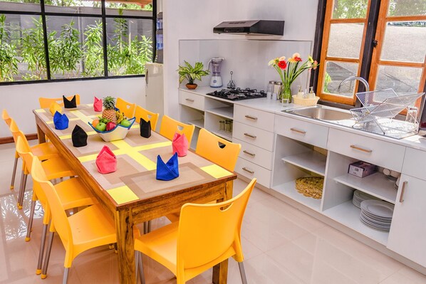 A modern kitchen with a dining area, featuring a wooden table, yellow chairs, a built-in stove, and a window with a view of greenery. - Pinnawala Backpack Residencies (Rambukkana)
