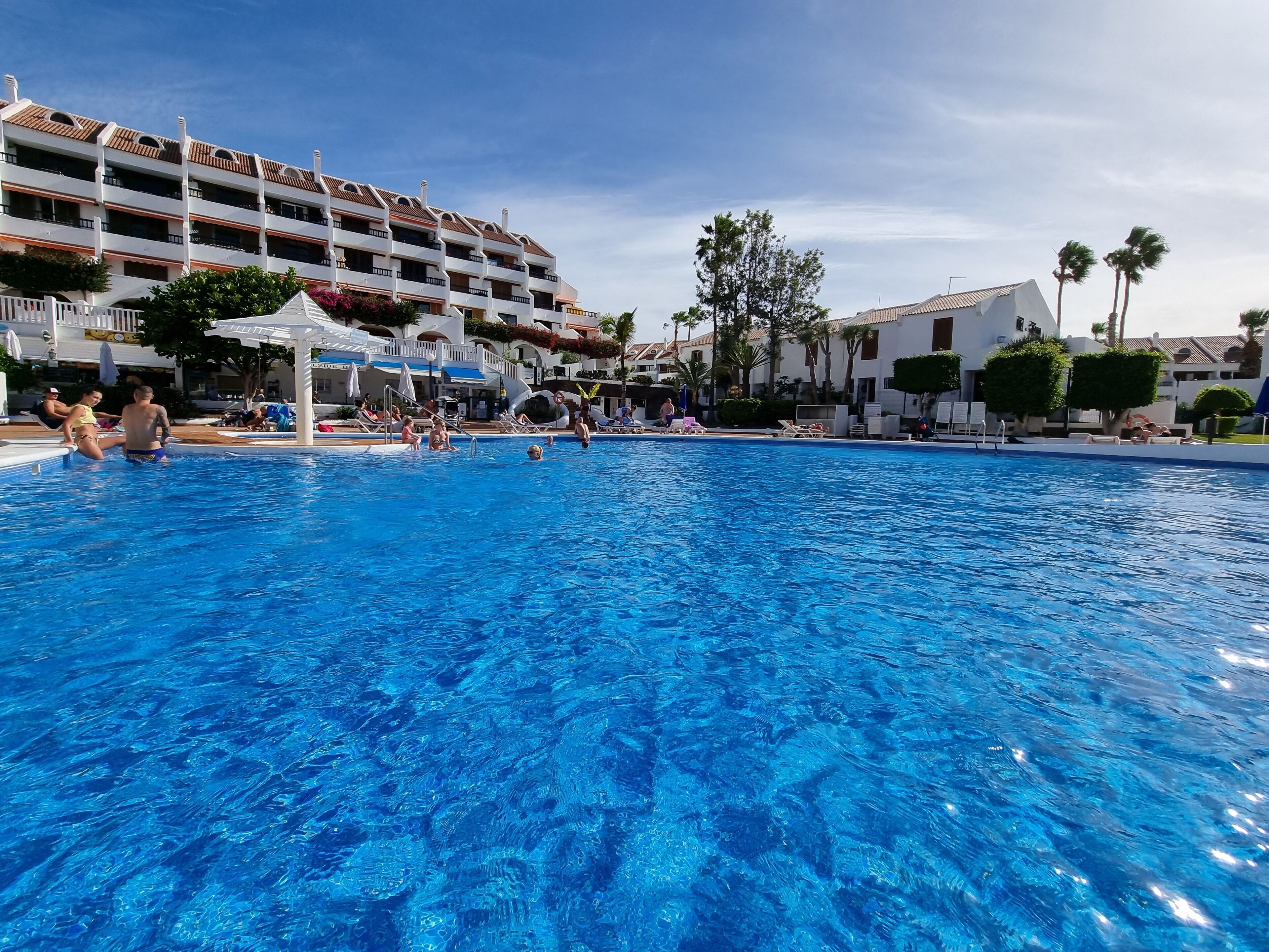 Uma piscina grande com pessoas ao redor, um edifício branco com varandas e palmeiras ao fundo.