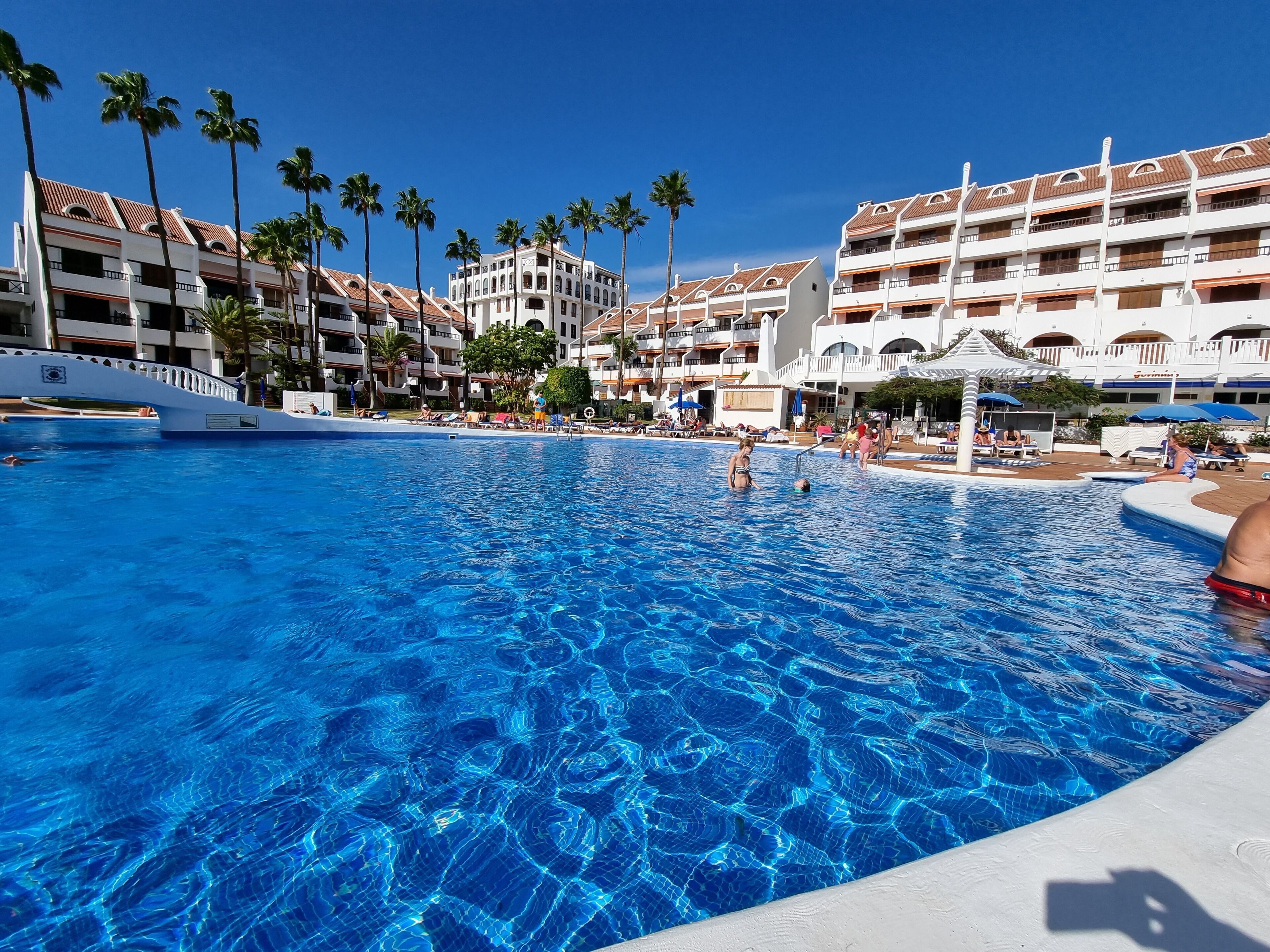 Een zwembad van een resort met helderblauw water, omgeven door palmbomen en meerlaagsgebouwen met balkons.