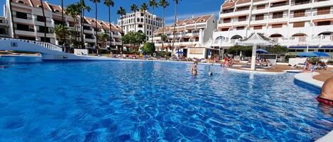 Une piscine d’un complexe hôtelier, avec une eau d’un bleu cristallin, entourée de palmiers et de bâtiments à plusieurs étages dotés de balcons.