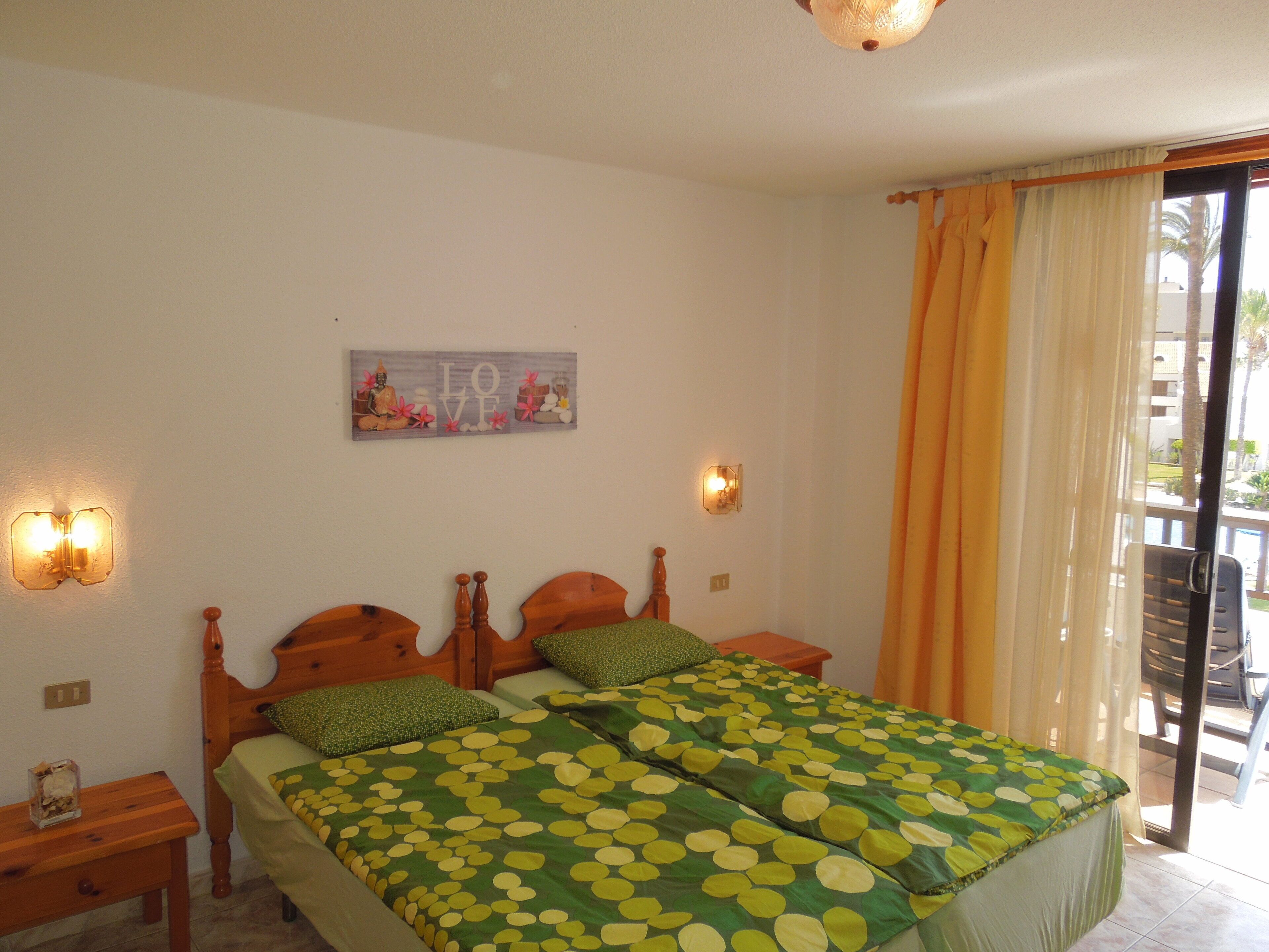 Un lit double avec une couvre-lit à pois verts et jaunes, deux tables de chevet, une lampe fixée au mur et une fenêtre avec des rideaux.