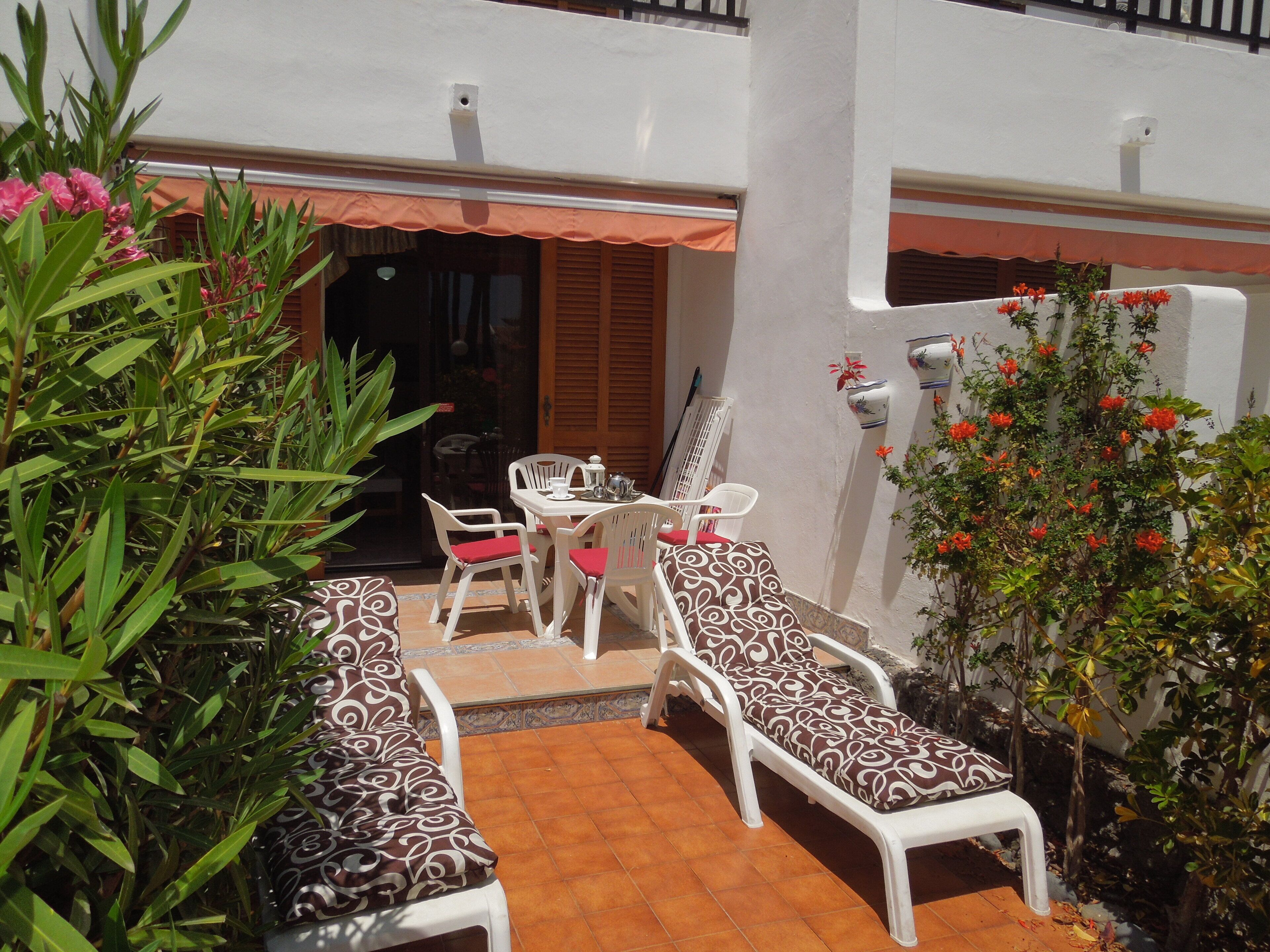 Eine Terrasse mit weißer Möbel, darunter ein Tisch und Stühle, umgeben von Pflanzen und einem kleinen Baum mit roten Blüten.