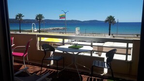 En balkon med bord og stole med udsigt over en strand, et flag, palmer og en klar himmel.