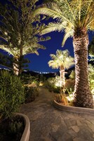 Un jardín nocturno con un sendero de piedra, palmeras y una pequeña estructura tipo tienda.