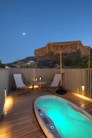 Una terraza de madera con un jacuzzi, dos sillones de descanso, una mesa con velas y un parasol de paja.