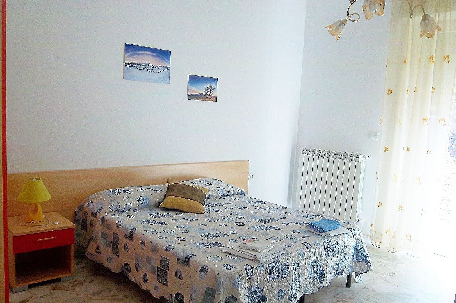 A bedroom with a bed, a nightstand with a lamp, and two wall-mounted pictures.
