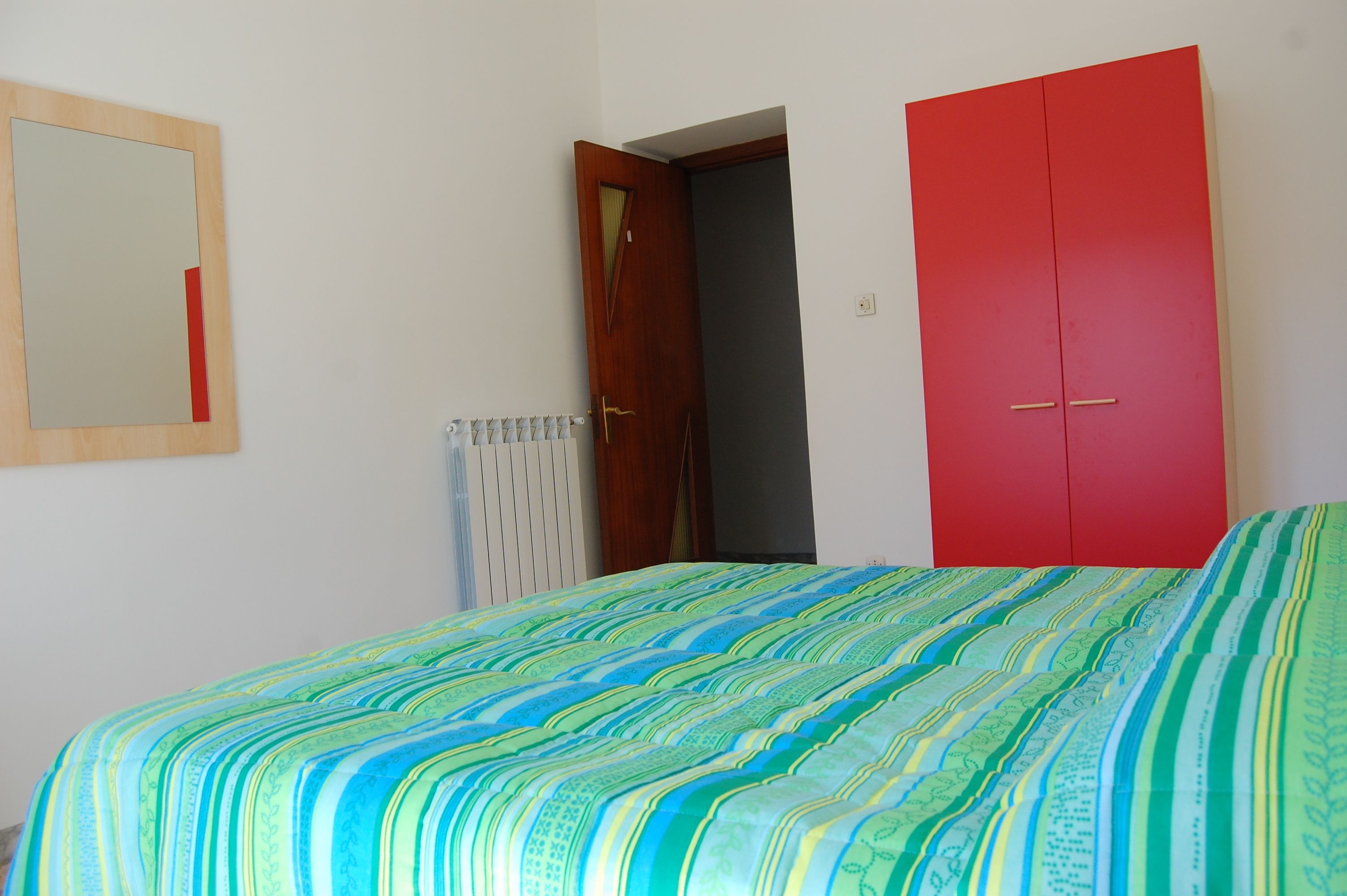 A bedroom with a bed, a red wardrobe, a mirror, and a door leading to another room.