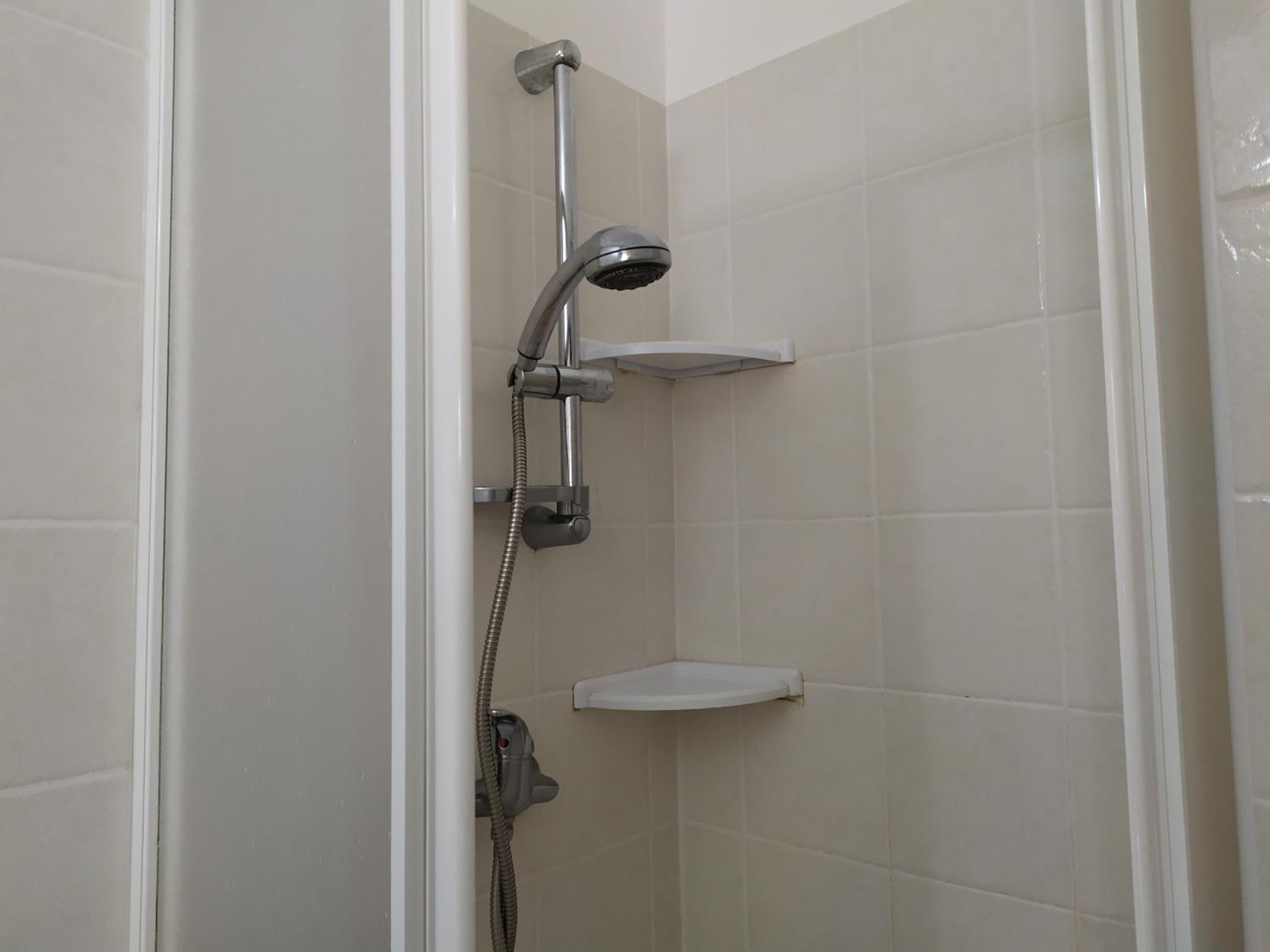 A shower with a handheld showerhead and a wall-mounted faucet, a small shelf, and tiled walls.