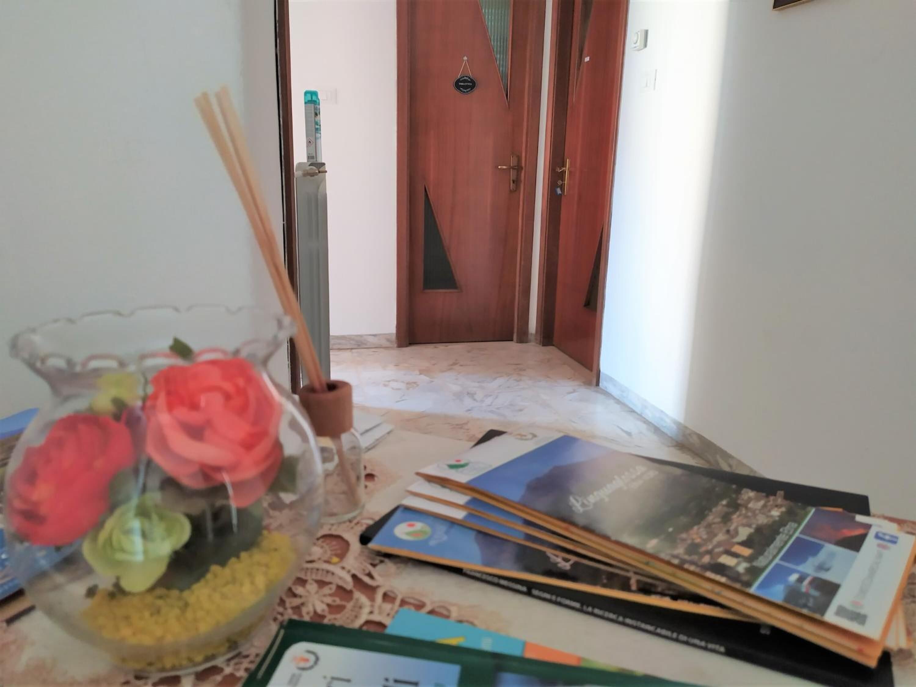 Una habitación con puerta de madera, una mesa con revistas y un florero de vidrio con flores.