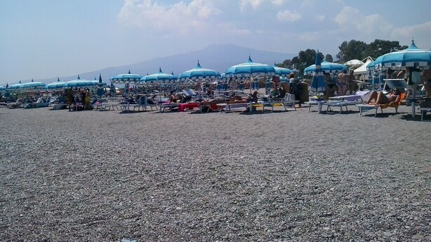 A beach with numerous umbrellas and sun loungers, a pebbled beach, and a mountain in the background. - Casa Valentina (Linguaglossa)