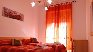 A bedroom with two beds, orange curtains, a painting on the wall, and a small table with towels. - Casa Valentina (Linguaglossa)