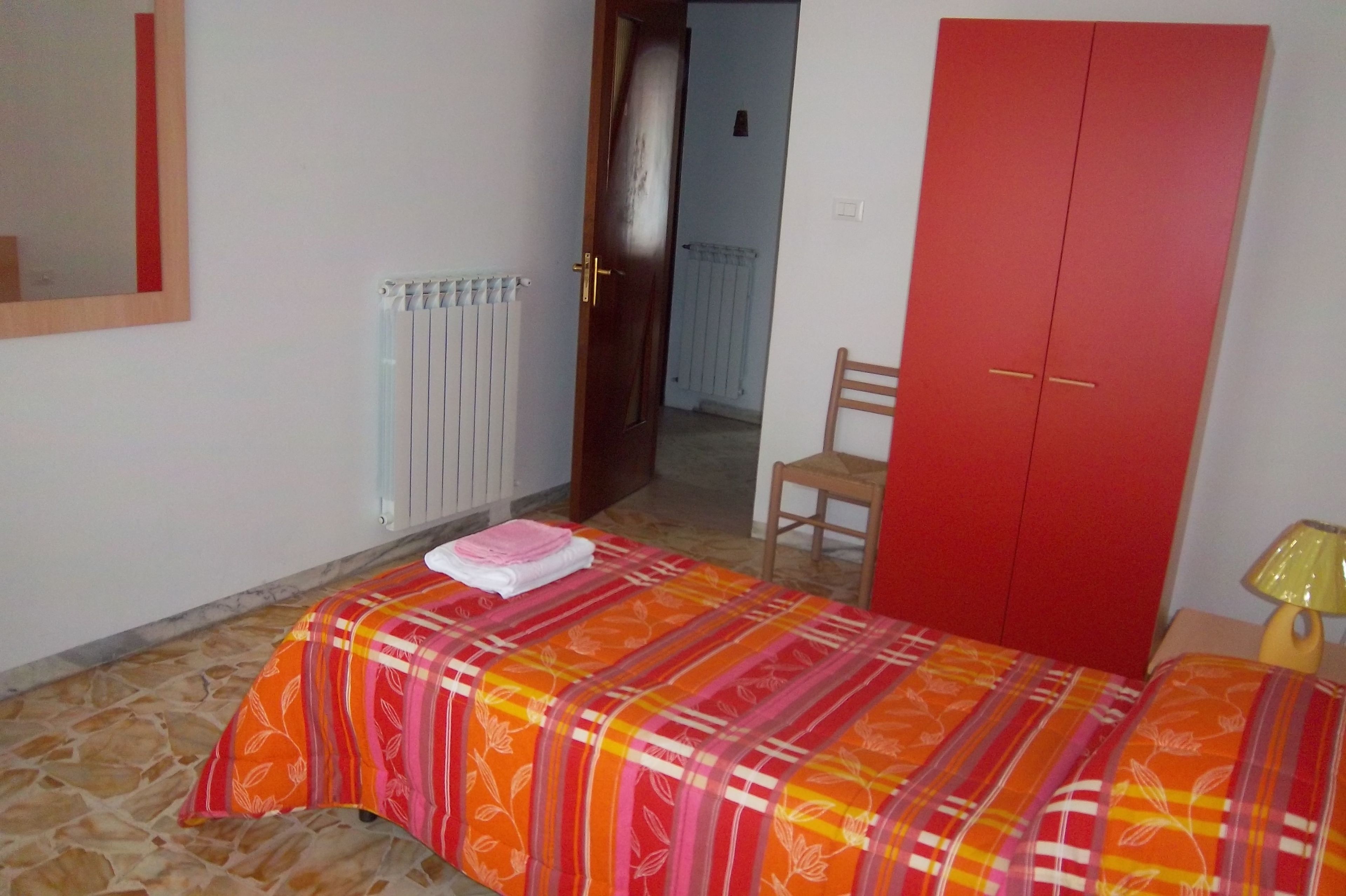 A bedroom with a bed, a red wardrobe, a chair, a radiator, and a mirror on the wall.